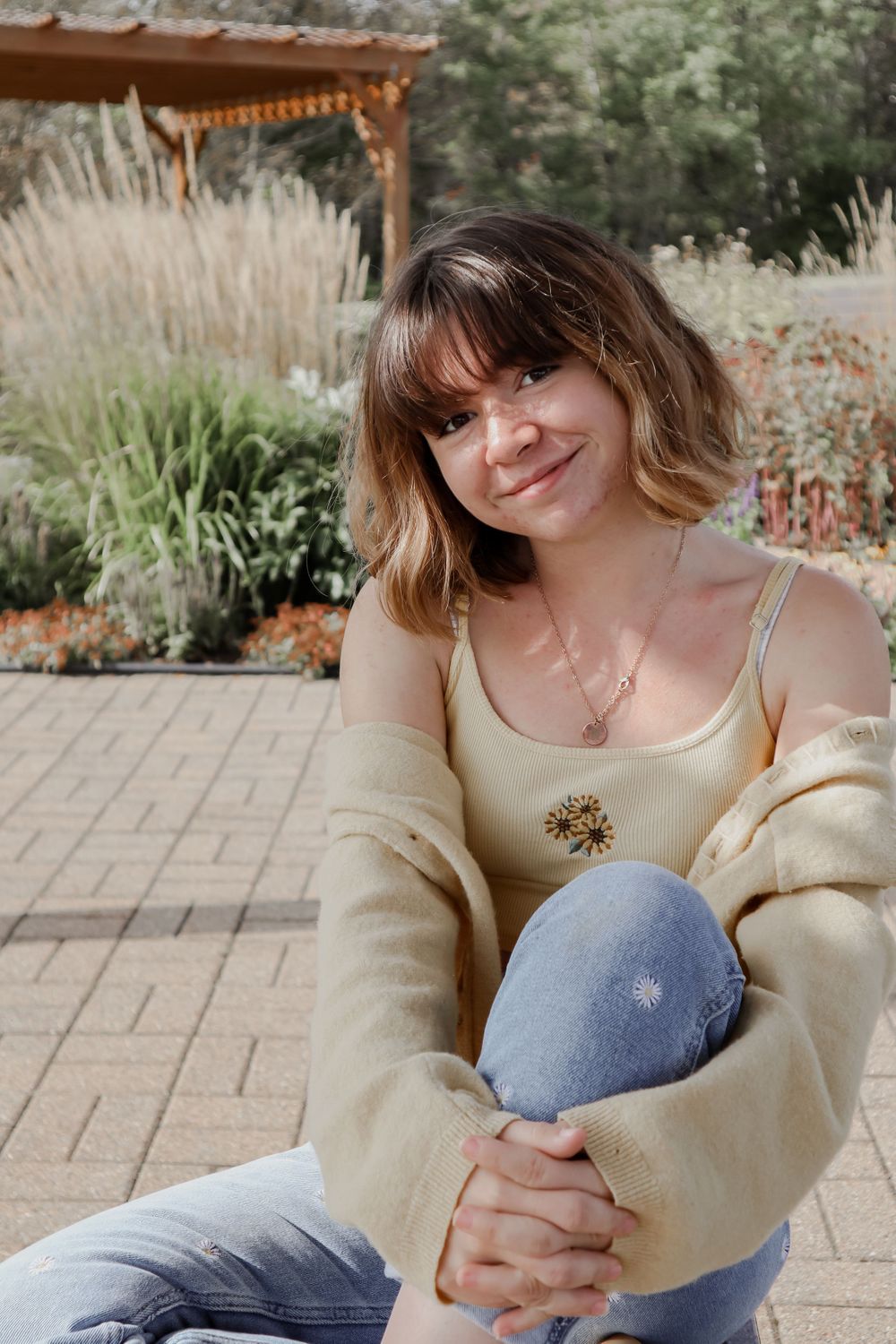 Person in a cream sweater and jeans sits on patio with ornamental grasses and pergola in background.