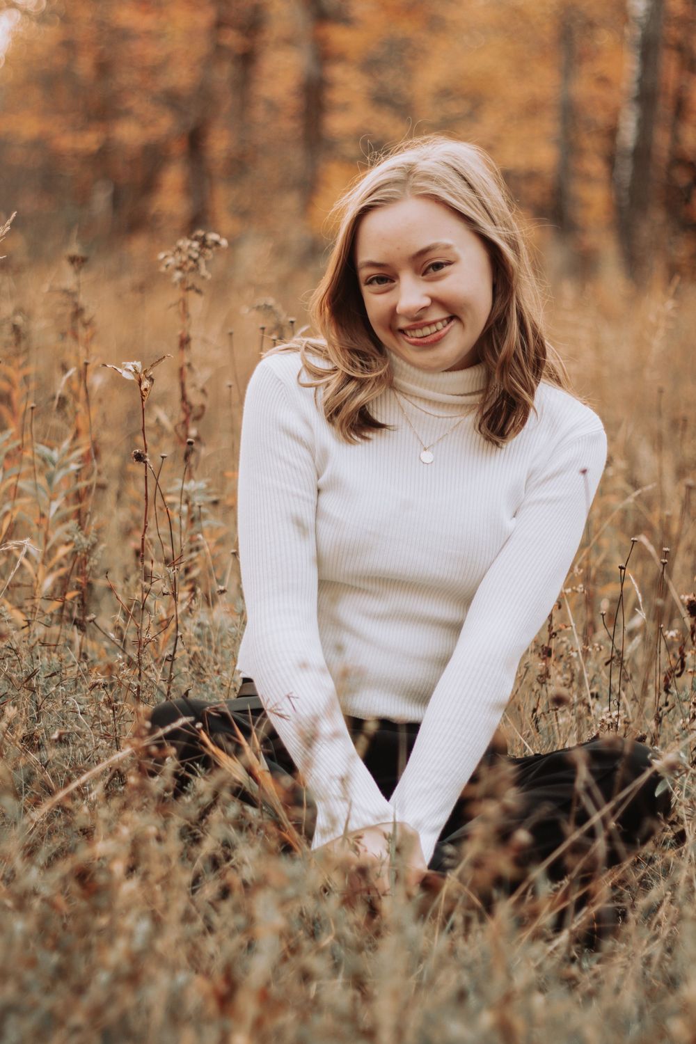Portrait of someone in a cream turtleneck sweater sitting among autumn wildflowers.