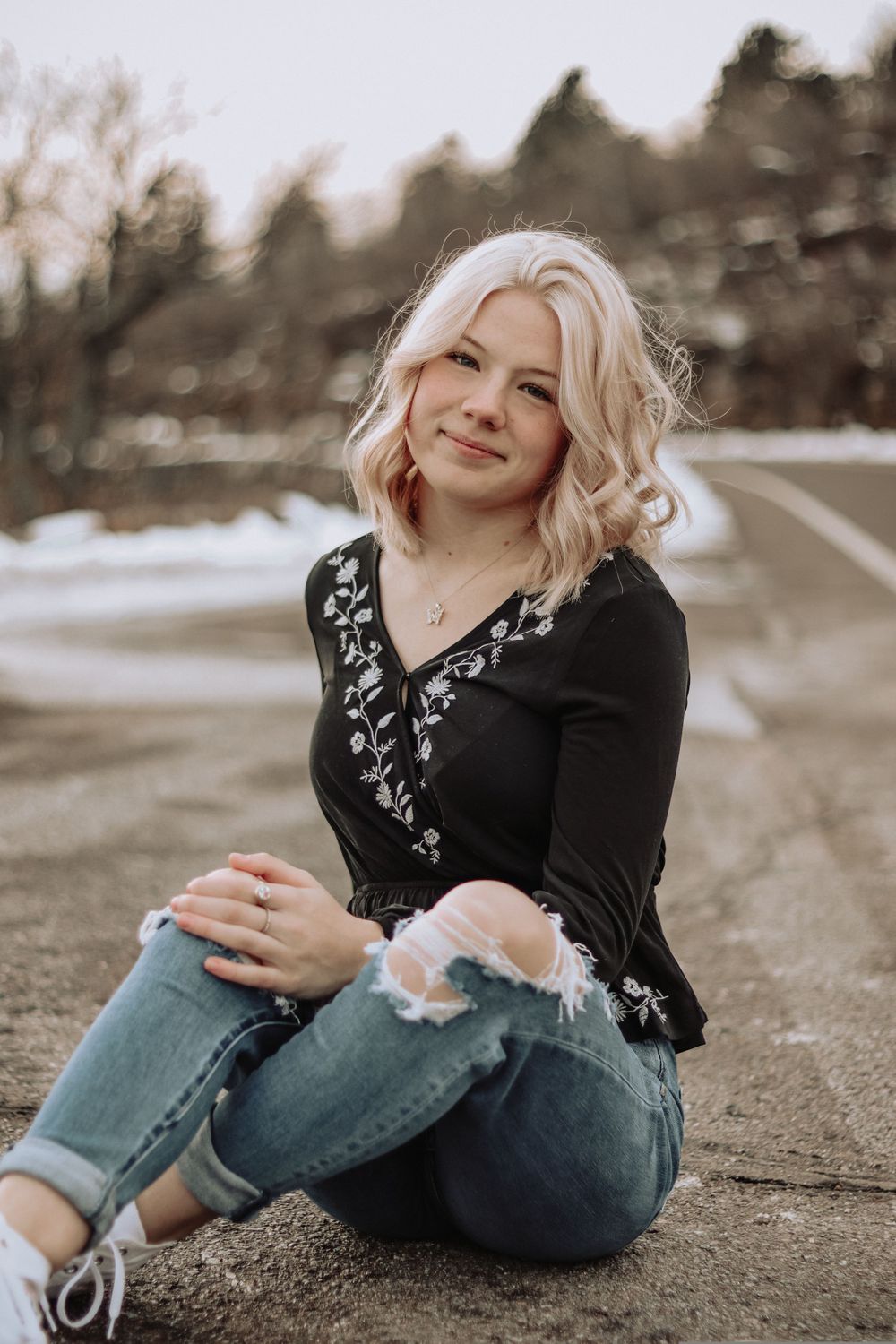 Casual outdoor photoshoot featuring someone in a black embroidered top and jeans sitting on the ground.