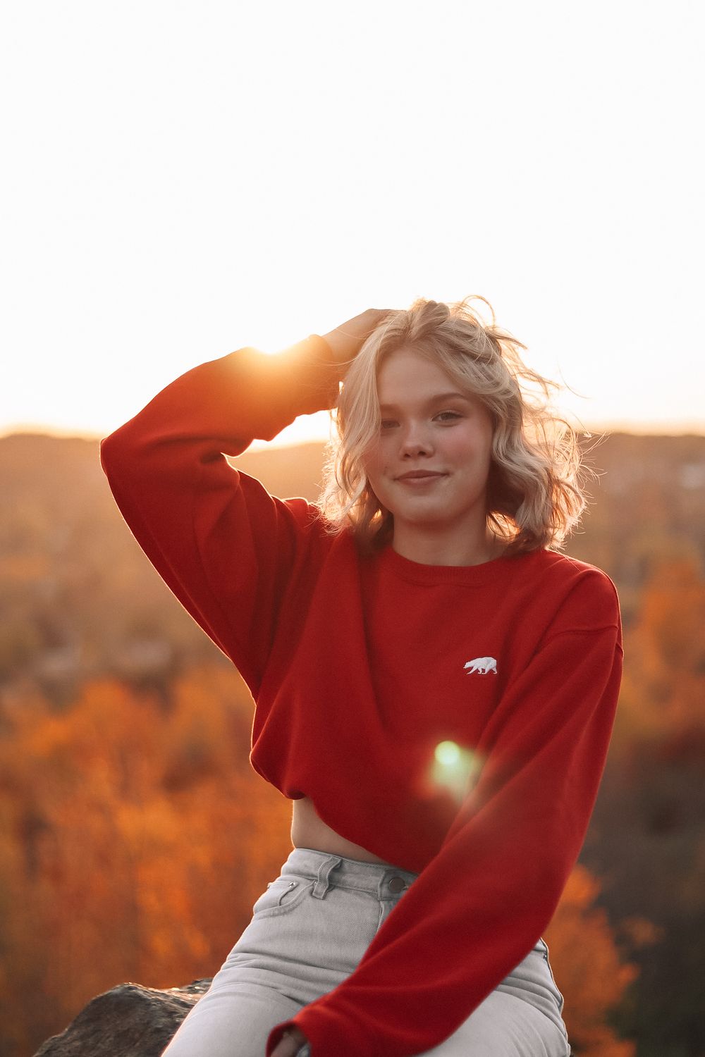 Person in red sweater poses outdoors during golden hour sunset with autumn colors in background.