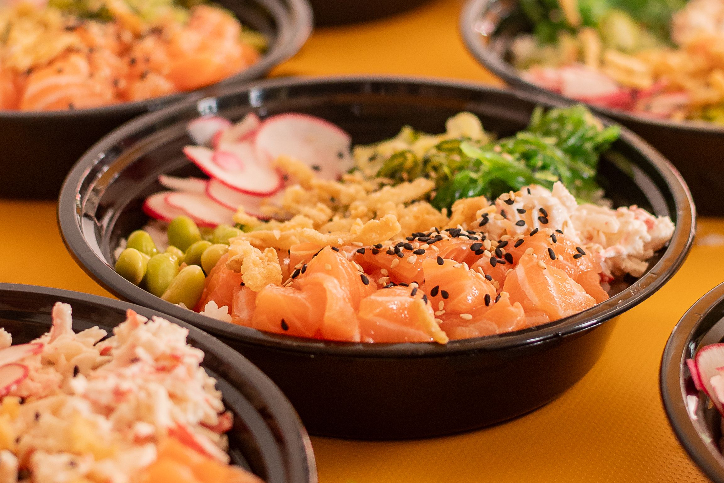 Multiple poke bowls with fresh salmon, vegetables and garnishes arranged on orange surface.
