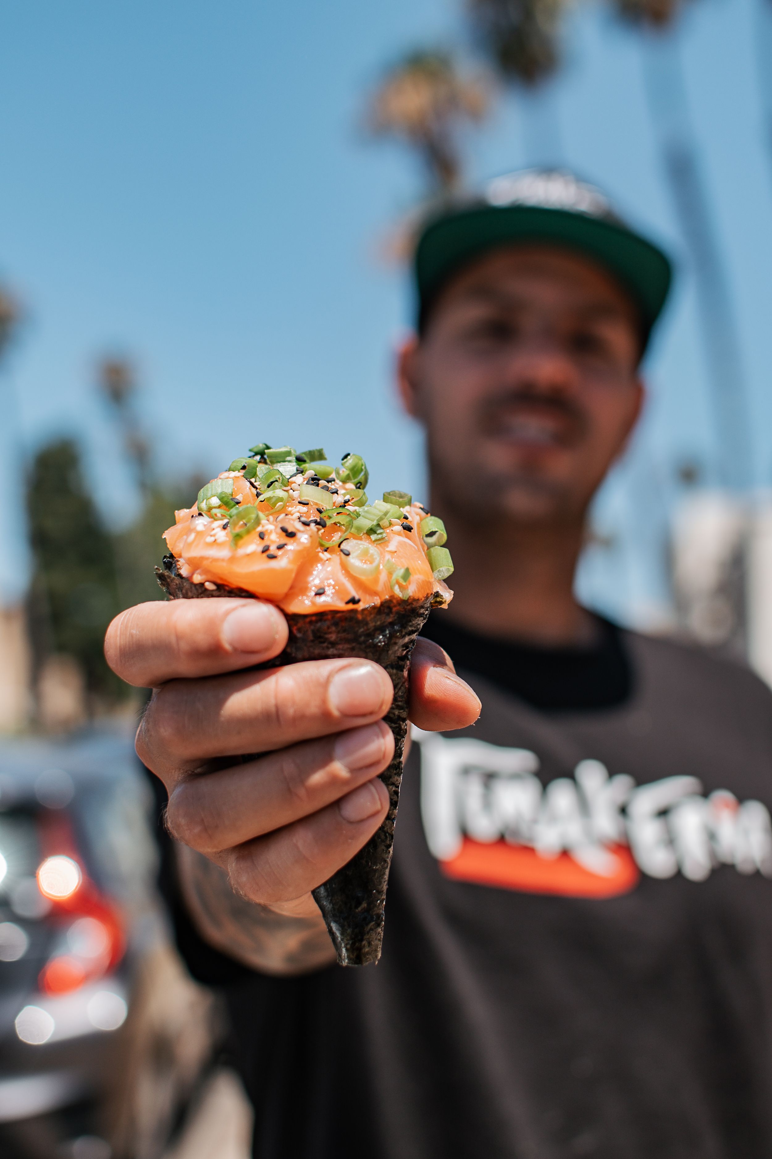 Hand holding a fresh sushi roll topped with sesame seeds and green garnish against a bright blue sky.