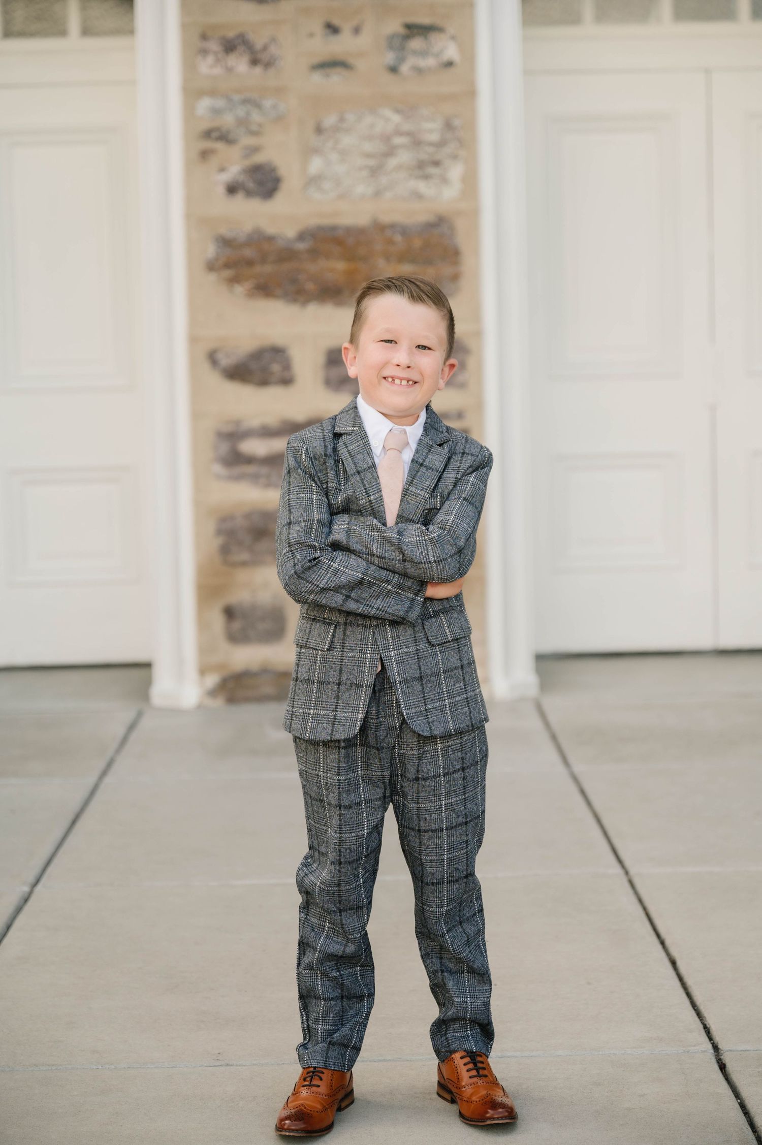 A young child in a gray plaid suit stands confidently in front of white garage doors.