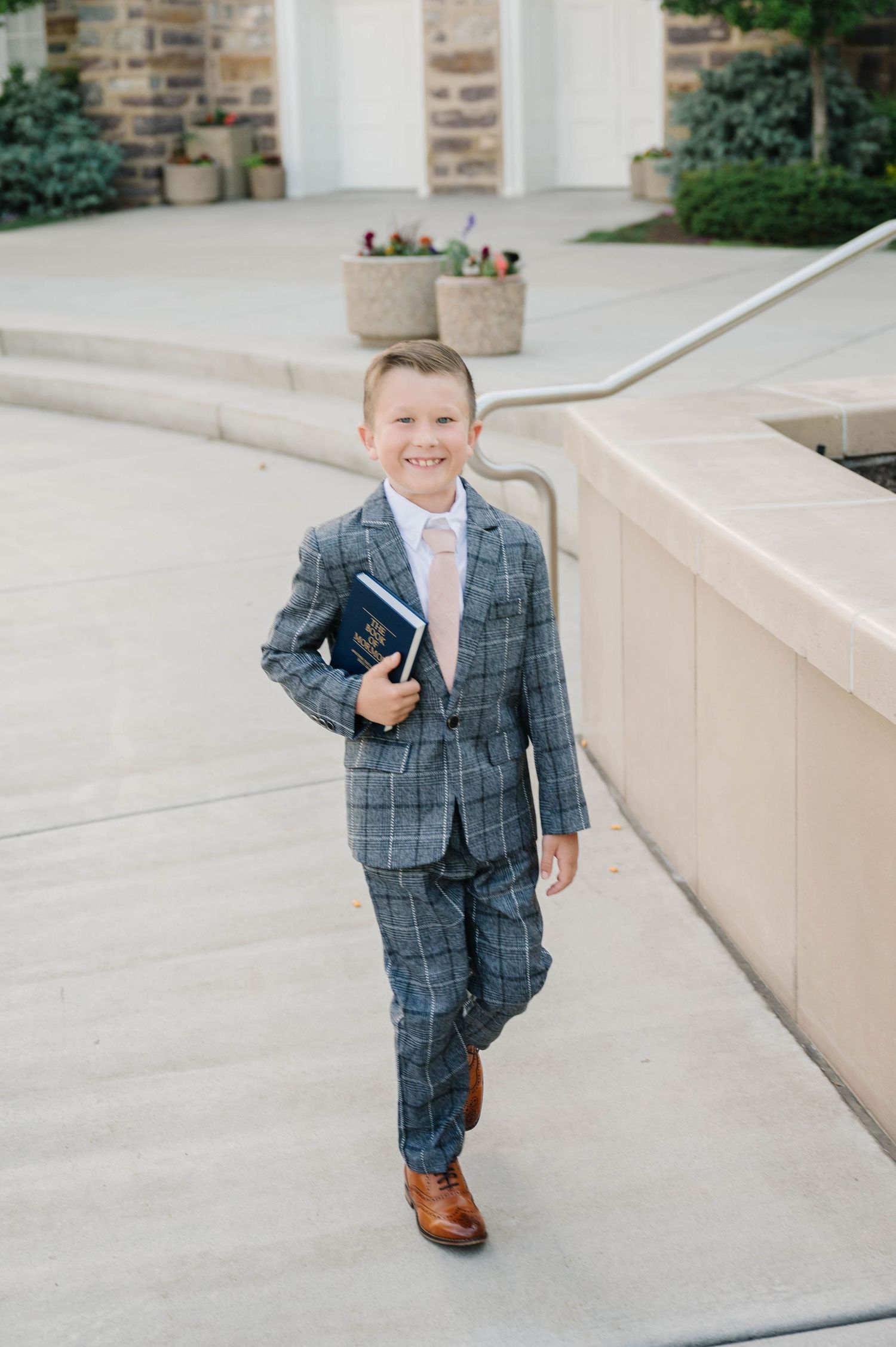 A young person in a grey plaid suit and pink tie holds a book while walking on a concrete path.