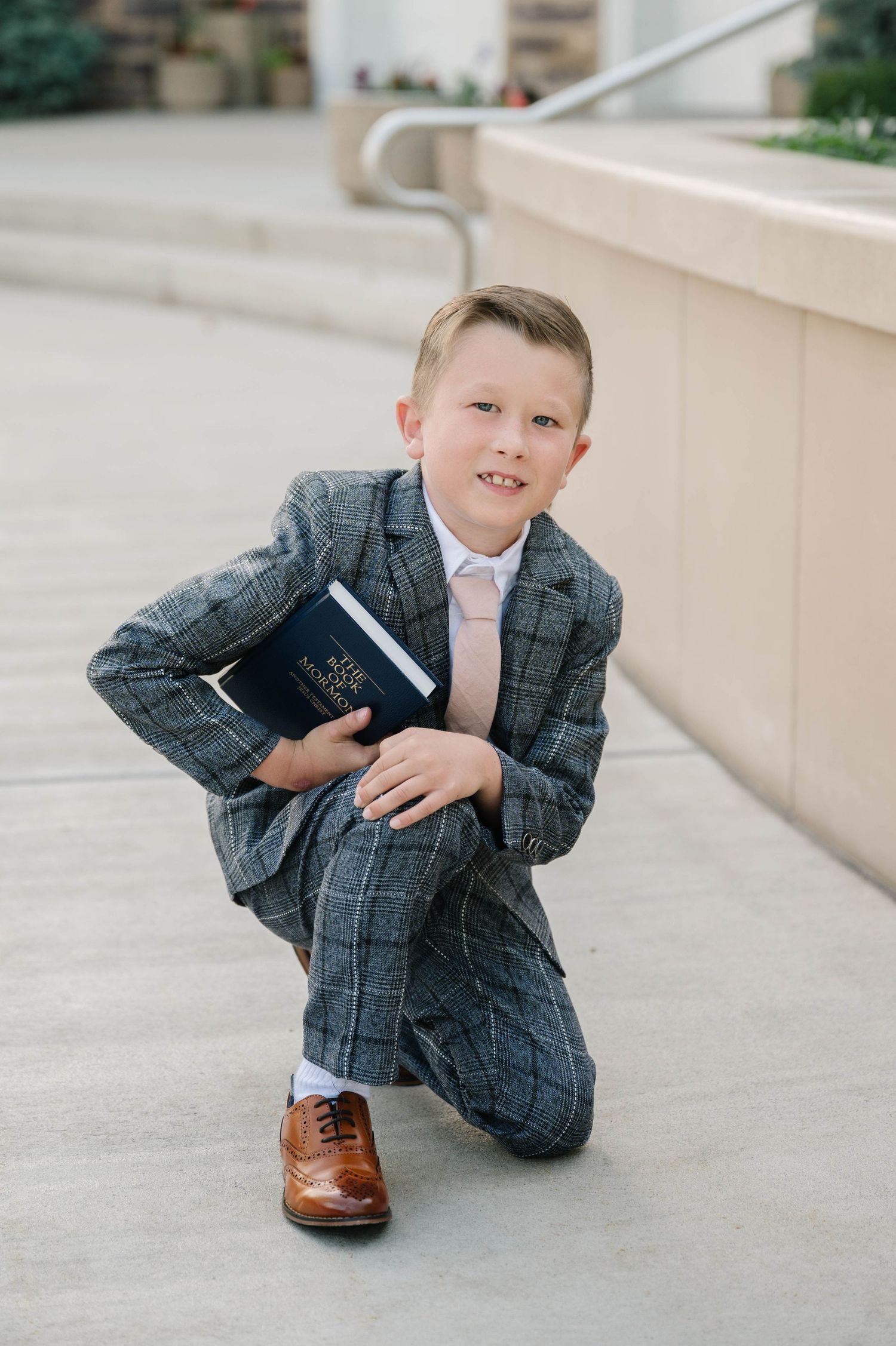 A person in a grey suit kneels on concrete while holding a book.