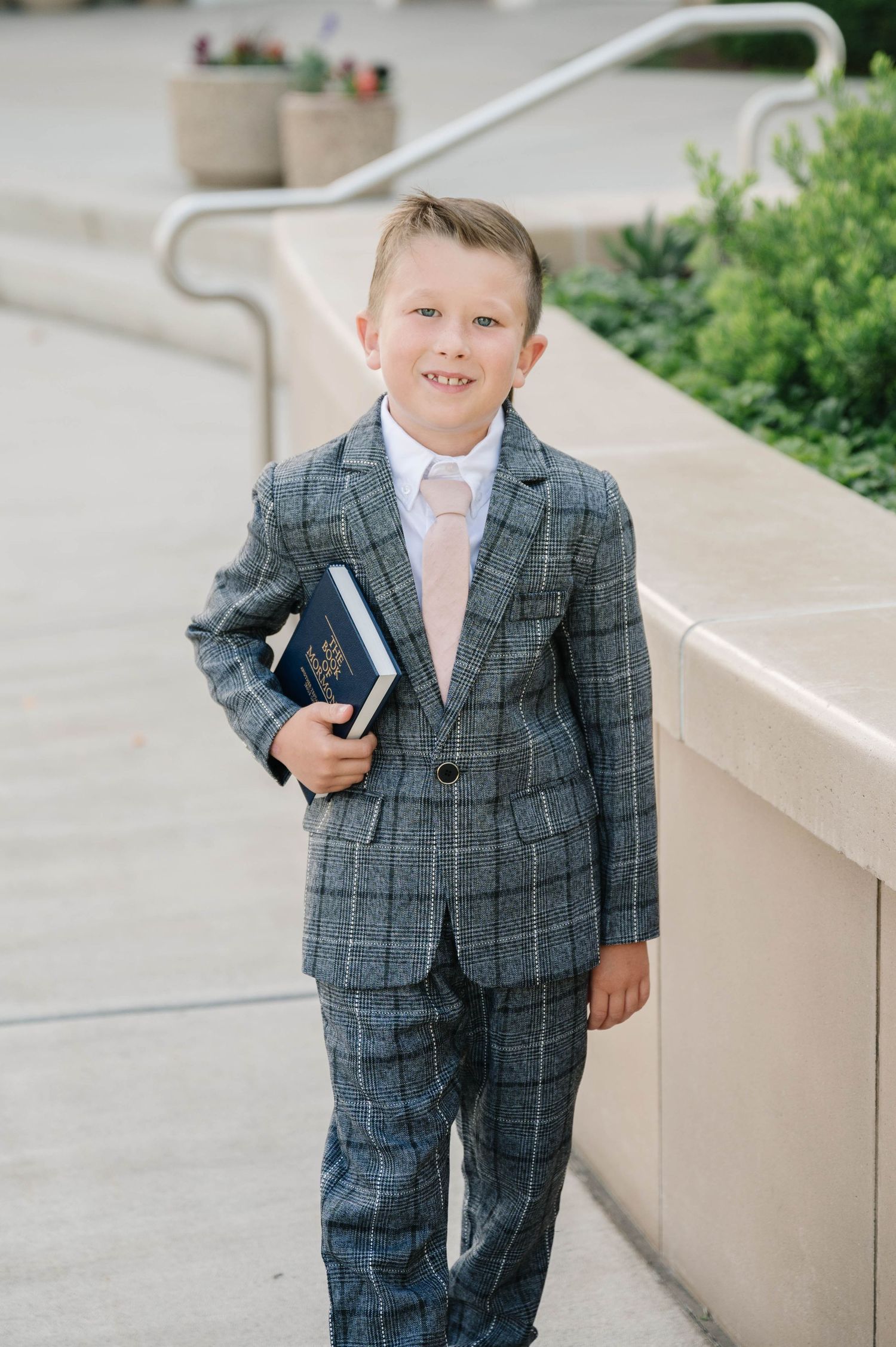 A person stands holding books while wearing a grey plaid suit and pink tie near concrete stairs.