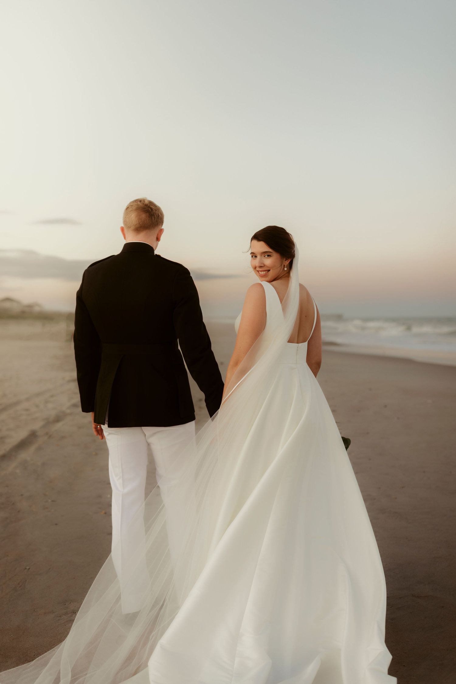 Couple walking on beach at sunset with bride in flowing white dress looking back at camera.