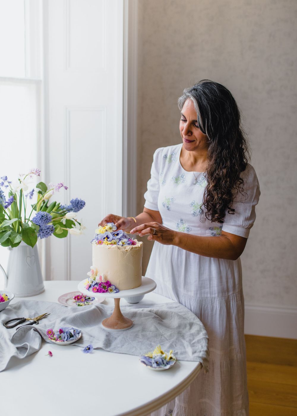 A person in white decorates a cake with colorful flowers on a marble table near windows with natural light.