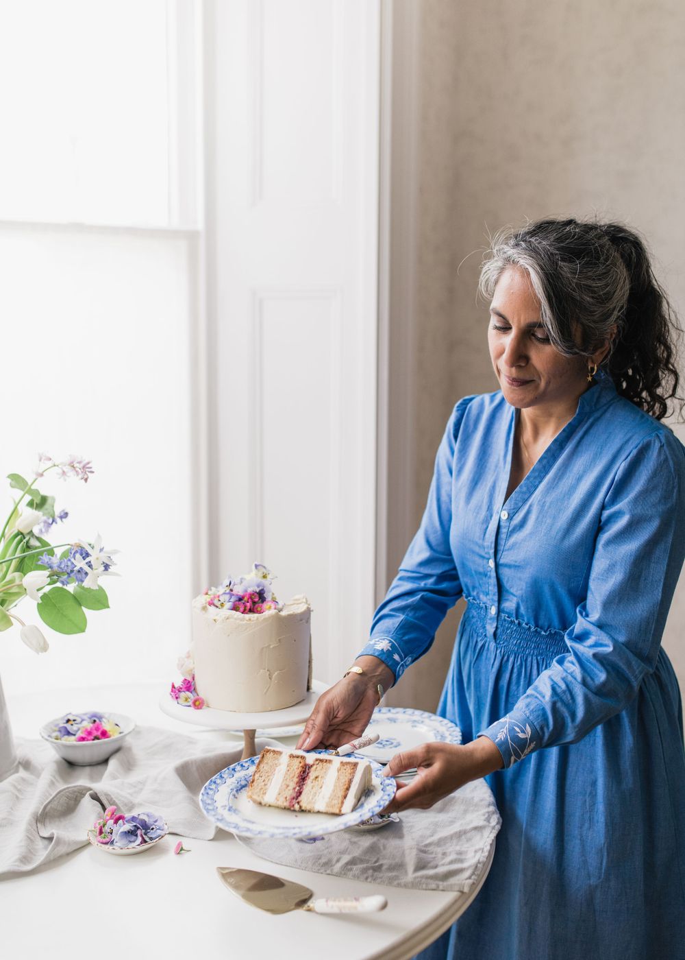 A person in a blue dress serves cake at a table decorated with purple flowers near a bright window.