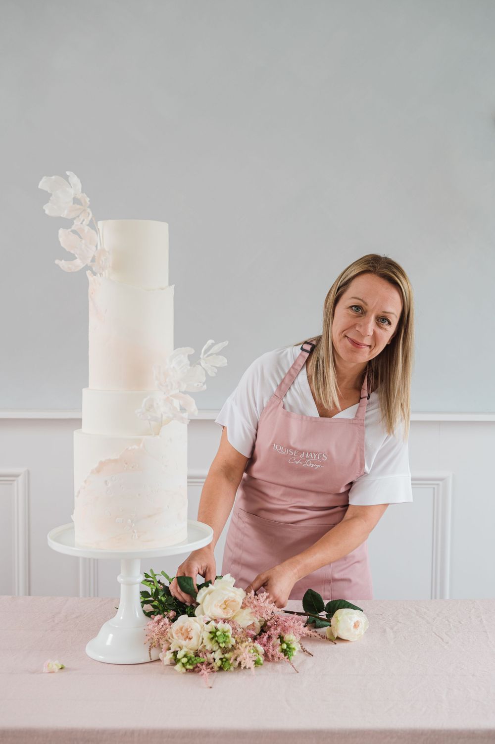 A wedding cake decorator arranges fresh pink and white flowers around an elegant white tiered cake.