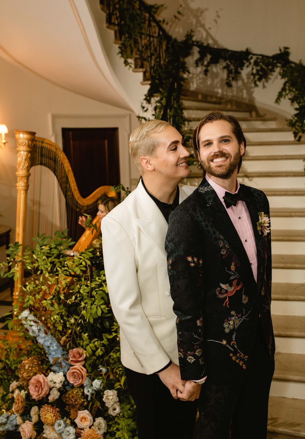 Elegant wedding couple posing by floral staircase with harp in background.