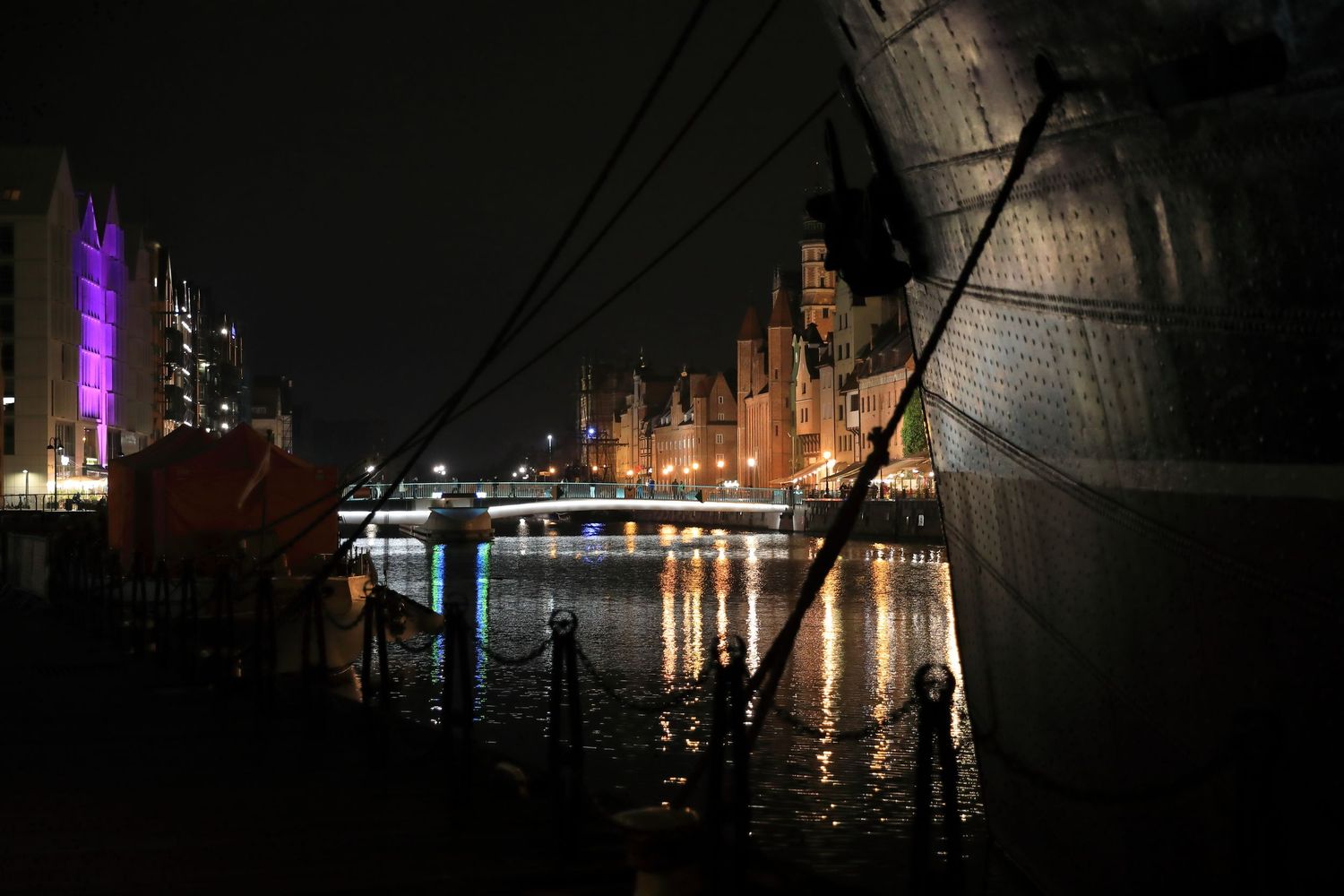 Night view of illuminated harbor buildings reflecting on dark water with moored ships and purple lighting.