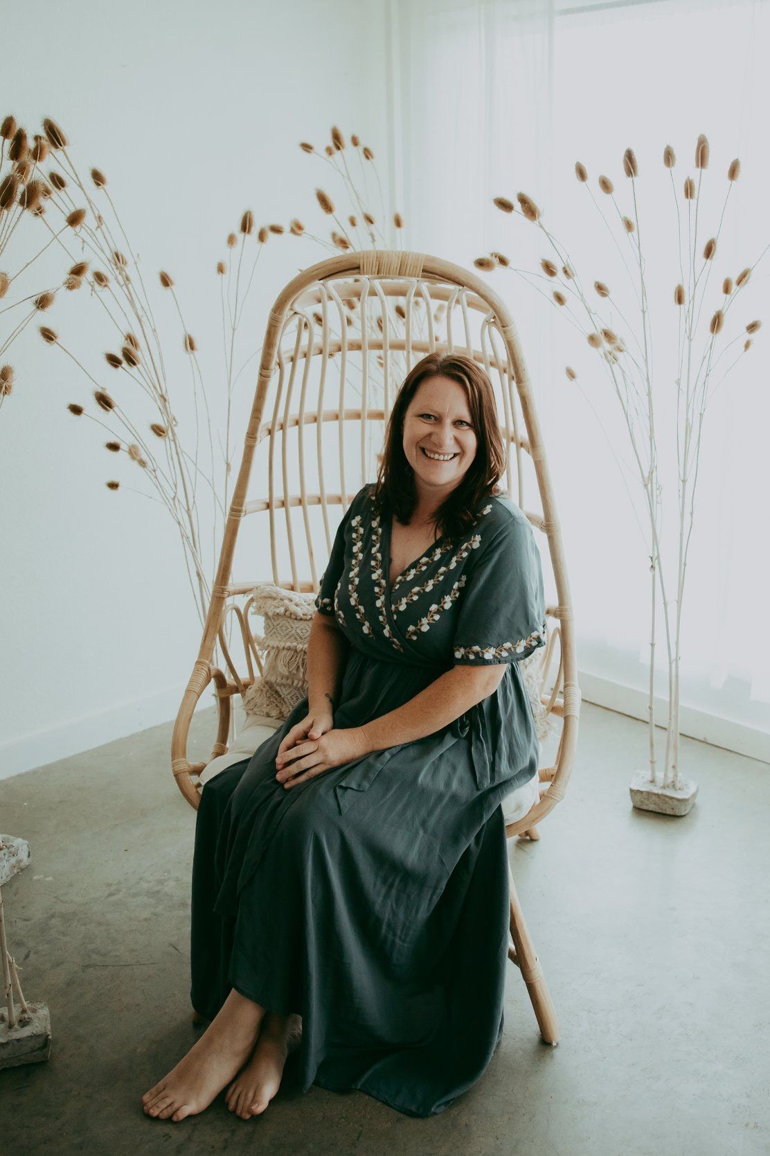 A person in a dark embroidered dress sits barefoot on a rattan hanging chair with dried grass decor.