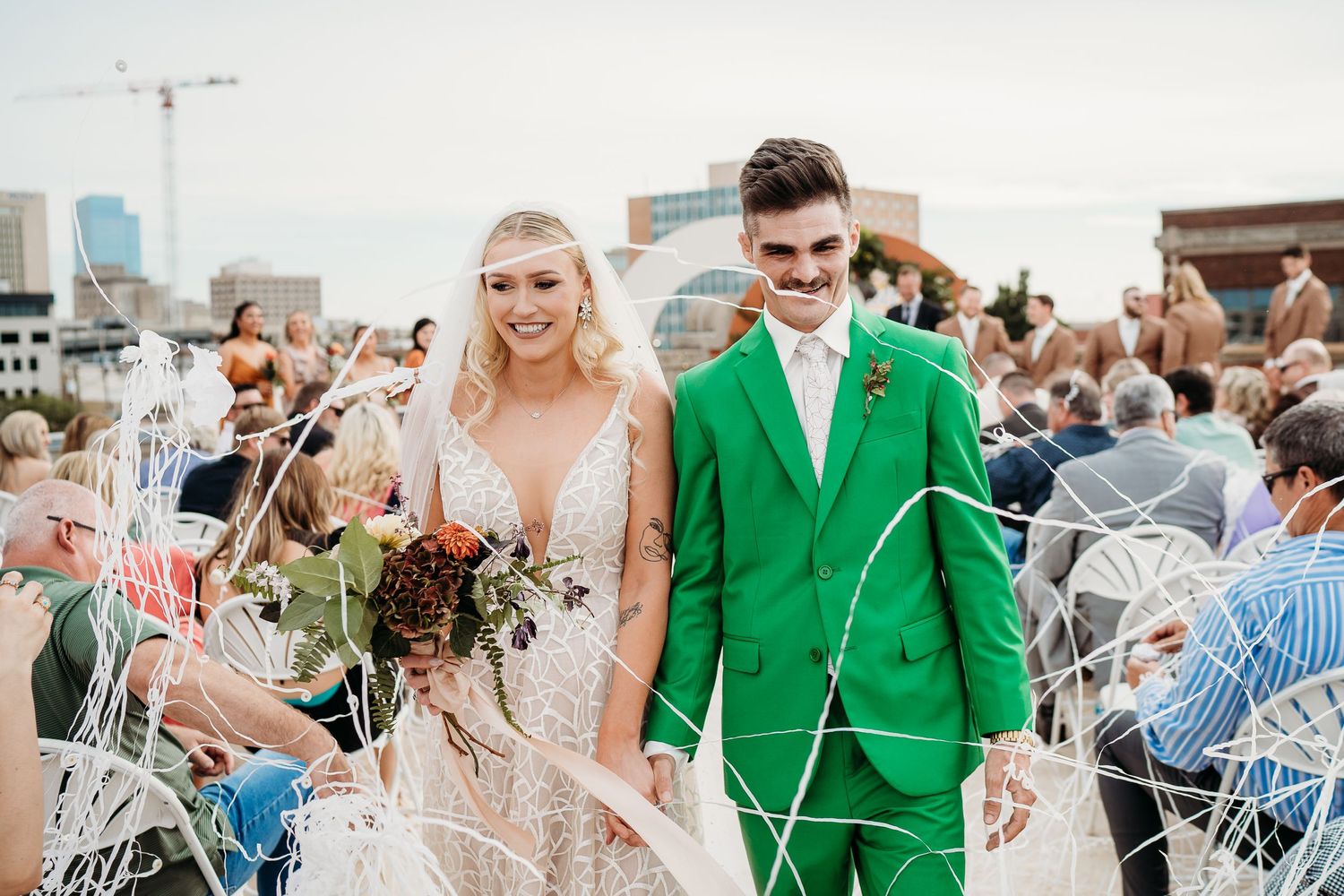 Wedding ceremony exit with guests on folding chairs as couple walks through in green suit and lace gown.