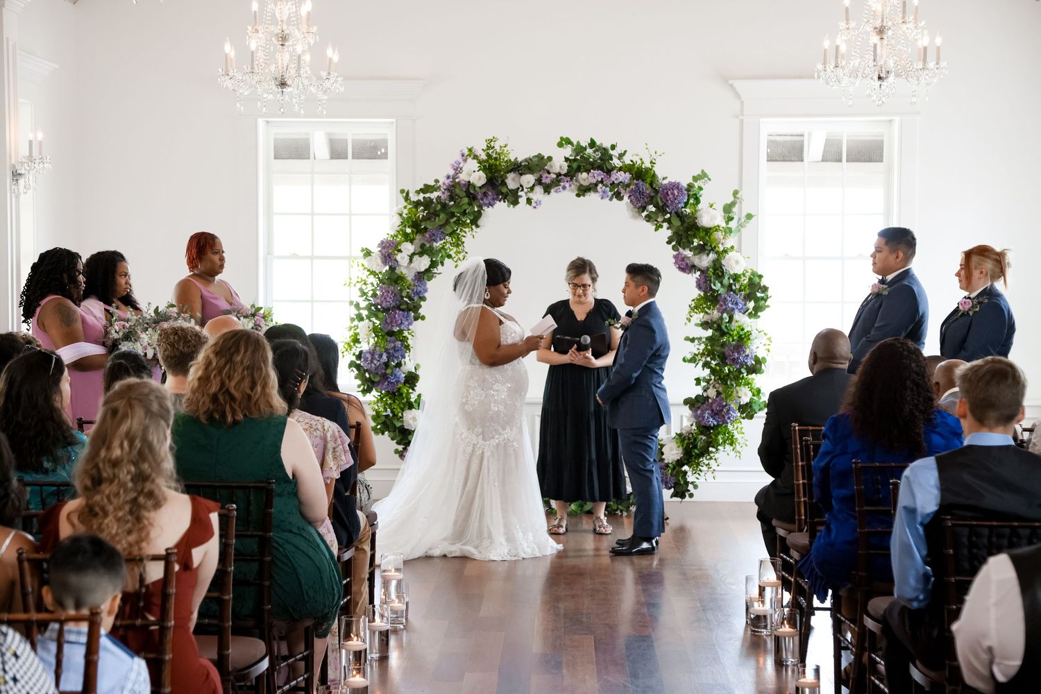 Wedding ceremony under floral arch in bright airy venue with white walls and chandelier.