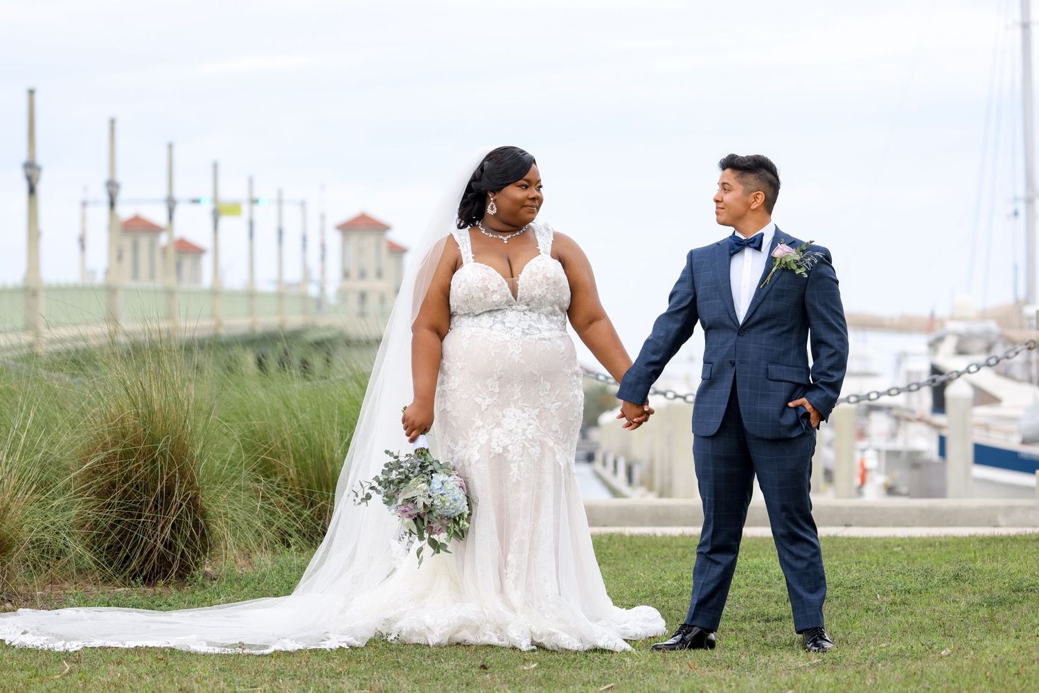 Couple holds hands near marina with bride in lace gown and groom in navy suit.