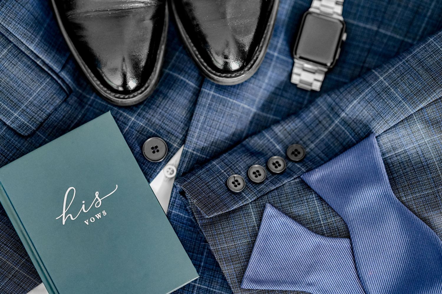 Groom's wedding accessories including black shoes, blue tie, and vow book on dark fabric.