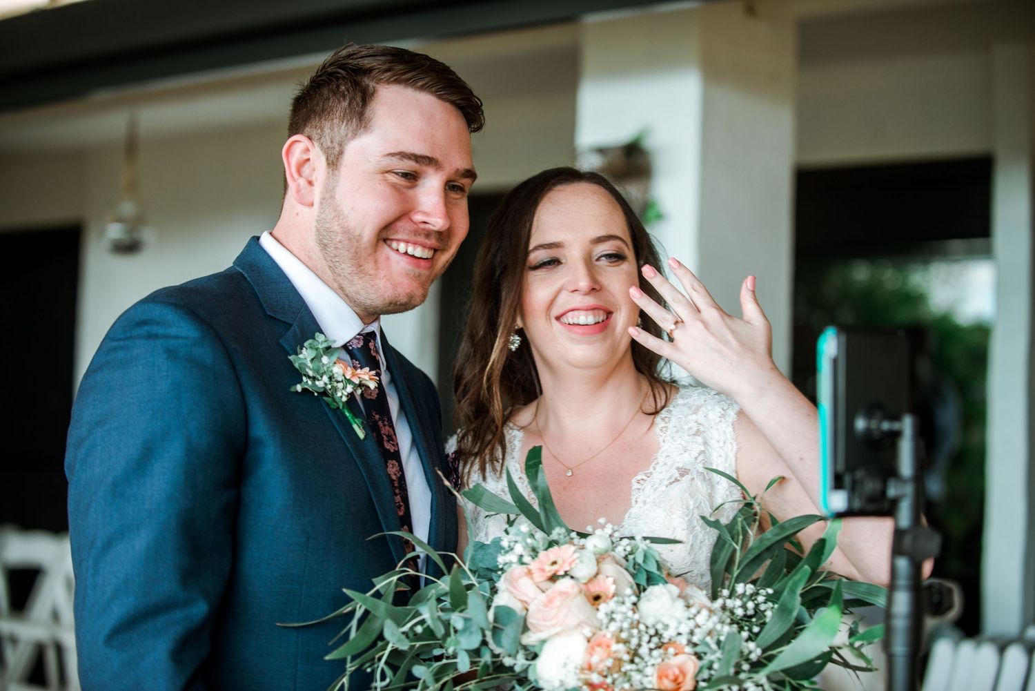 A joyful wedding couple smiles while holding a lush green and peach floral bouquet.