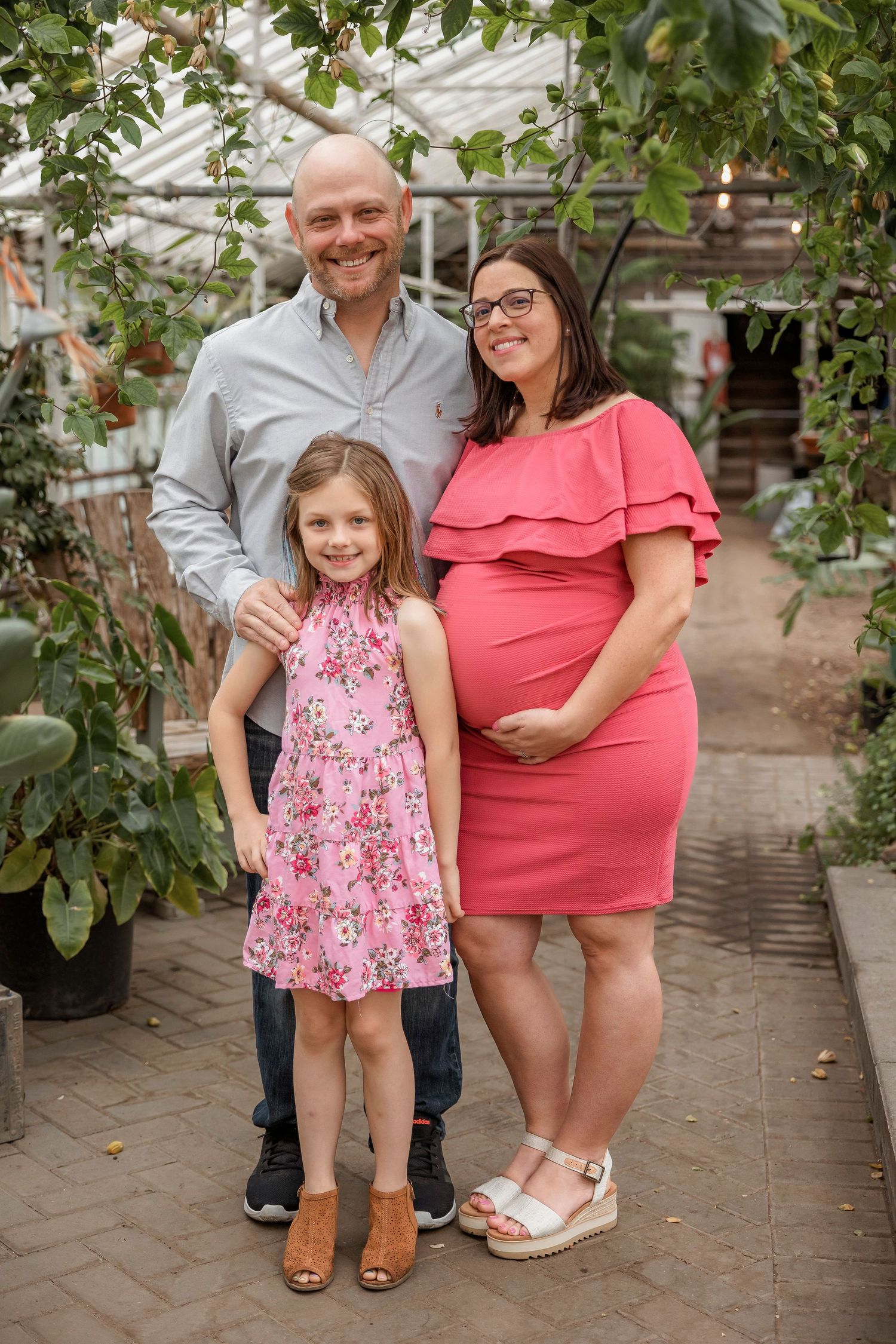 A family poses together in a greenhouse, with a young girl in a pink floral dress and expecting mother in coral dress.