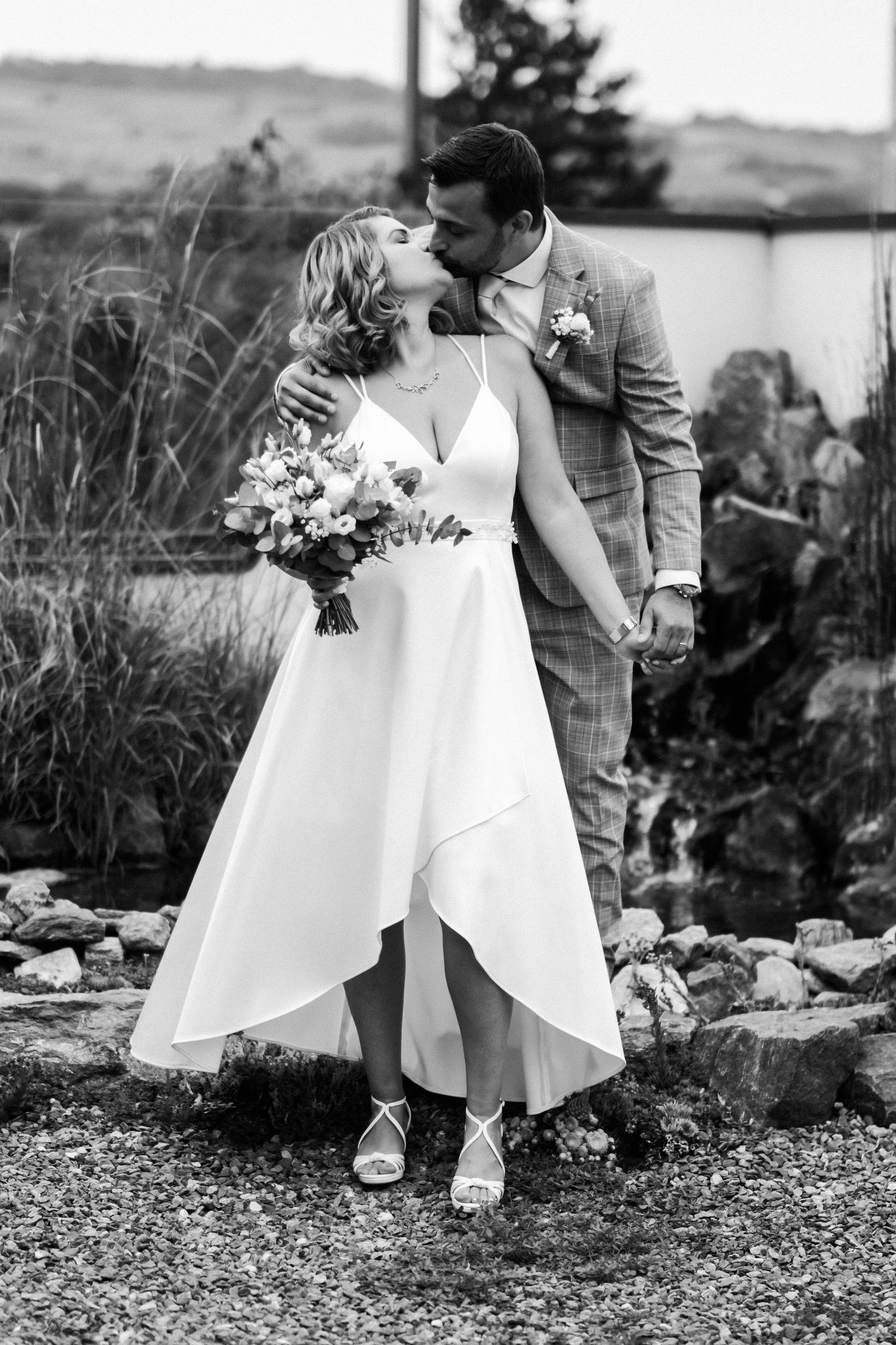 Black and white wedding photo of newlyweds sharing romantic kiss outdoors.