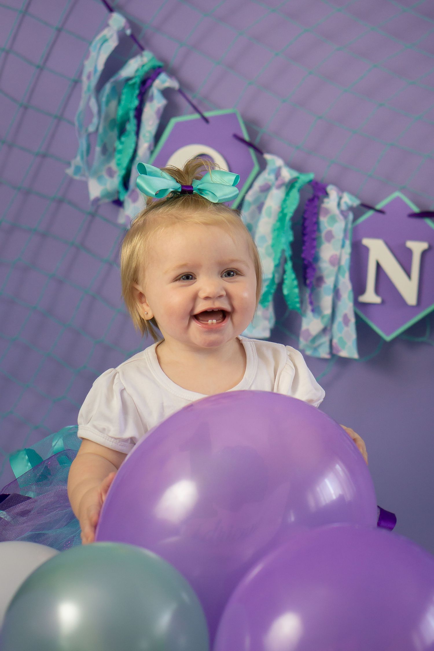 Happy toddler surrounded by purple and teal balloons at a birthday party.