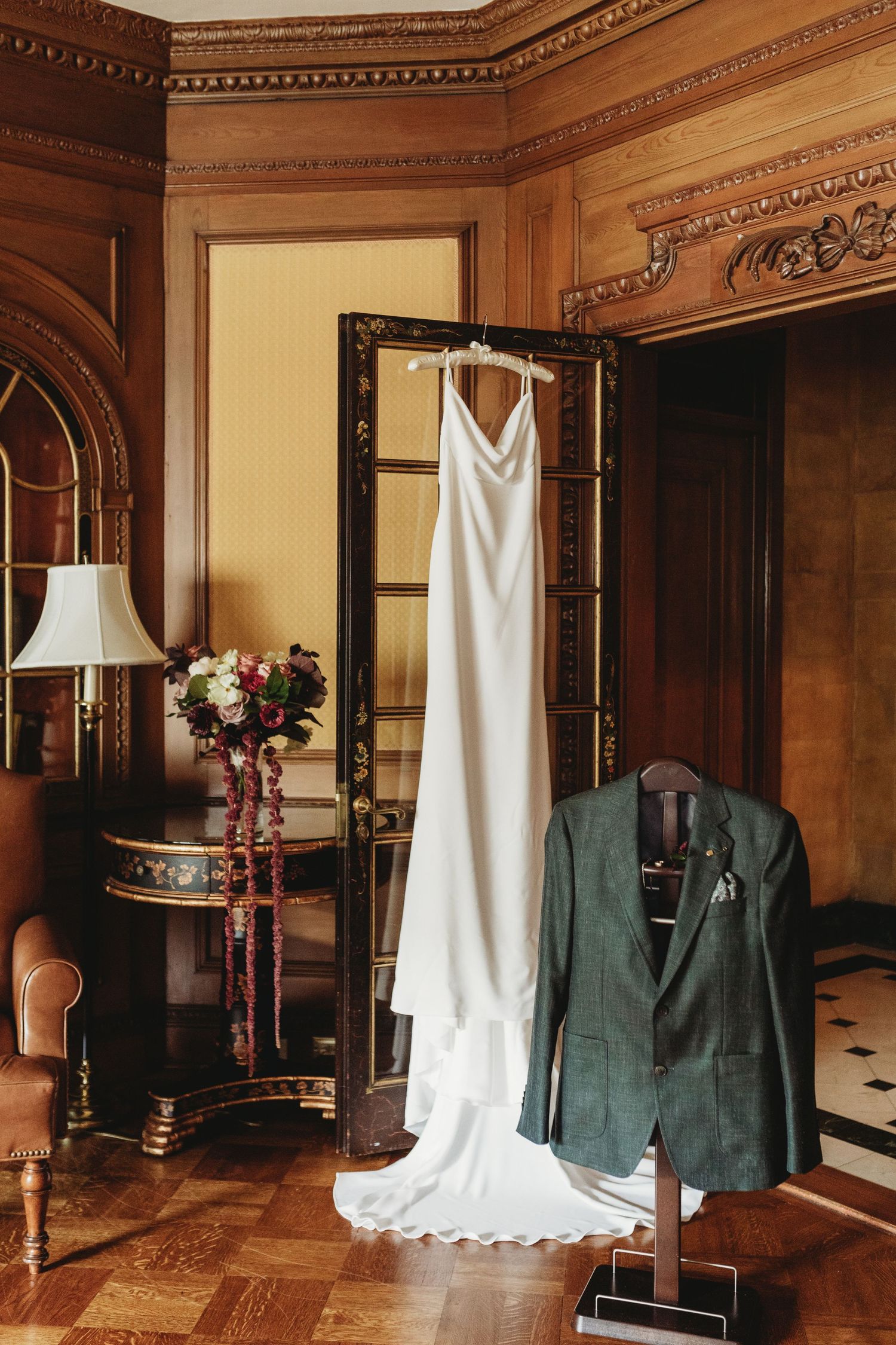A white wedding dress and green suit jacket hanging in an elegant wood-paneled room with ornate details.