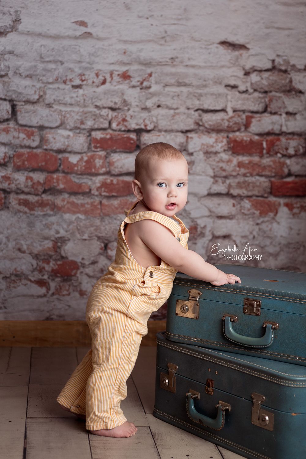 Standing baby boy wearing vintage overalls with vintage suitcases, brick wall.  Wisconsin Baby Portrait