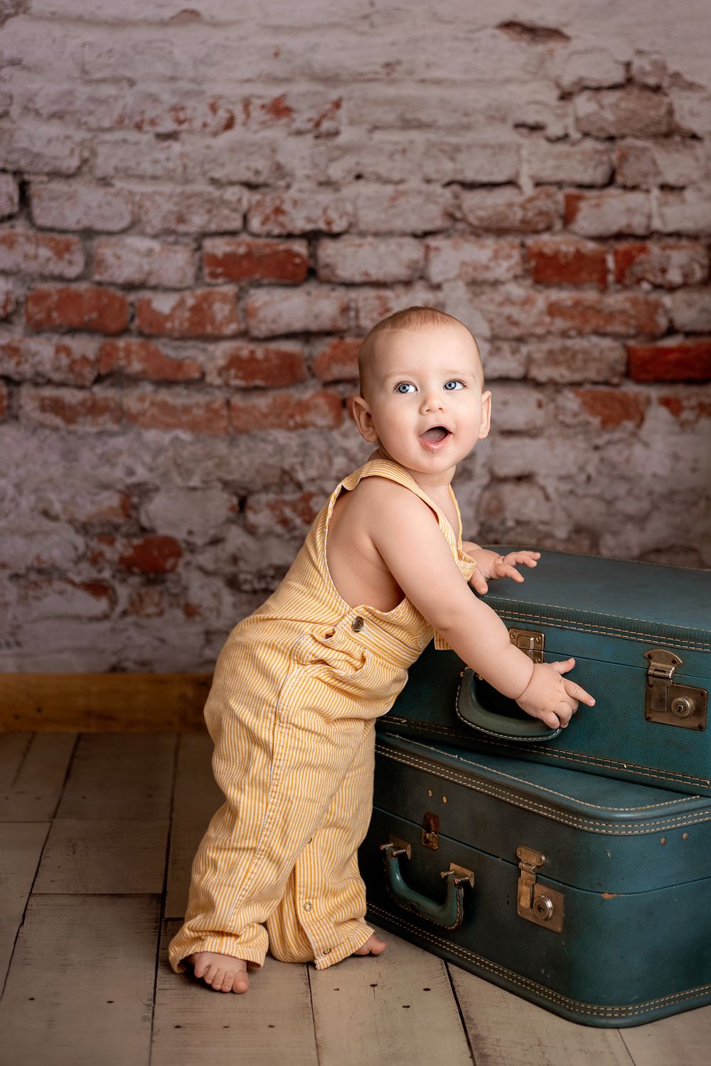 Standing baby boy wearing vintage overalls with vintage suitcases, brick wall.  Wisconsin Baby Portrait