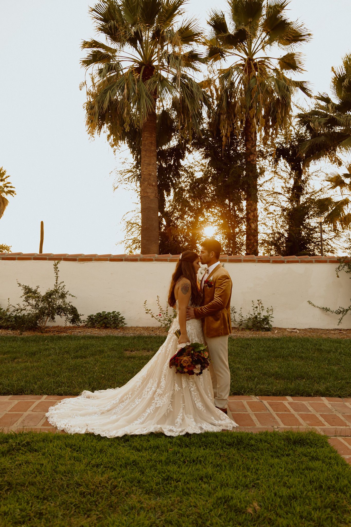 Sunset wedding portrait with palm trees and long white dress against Spanish-style wall.