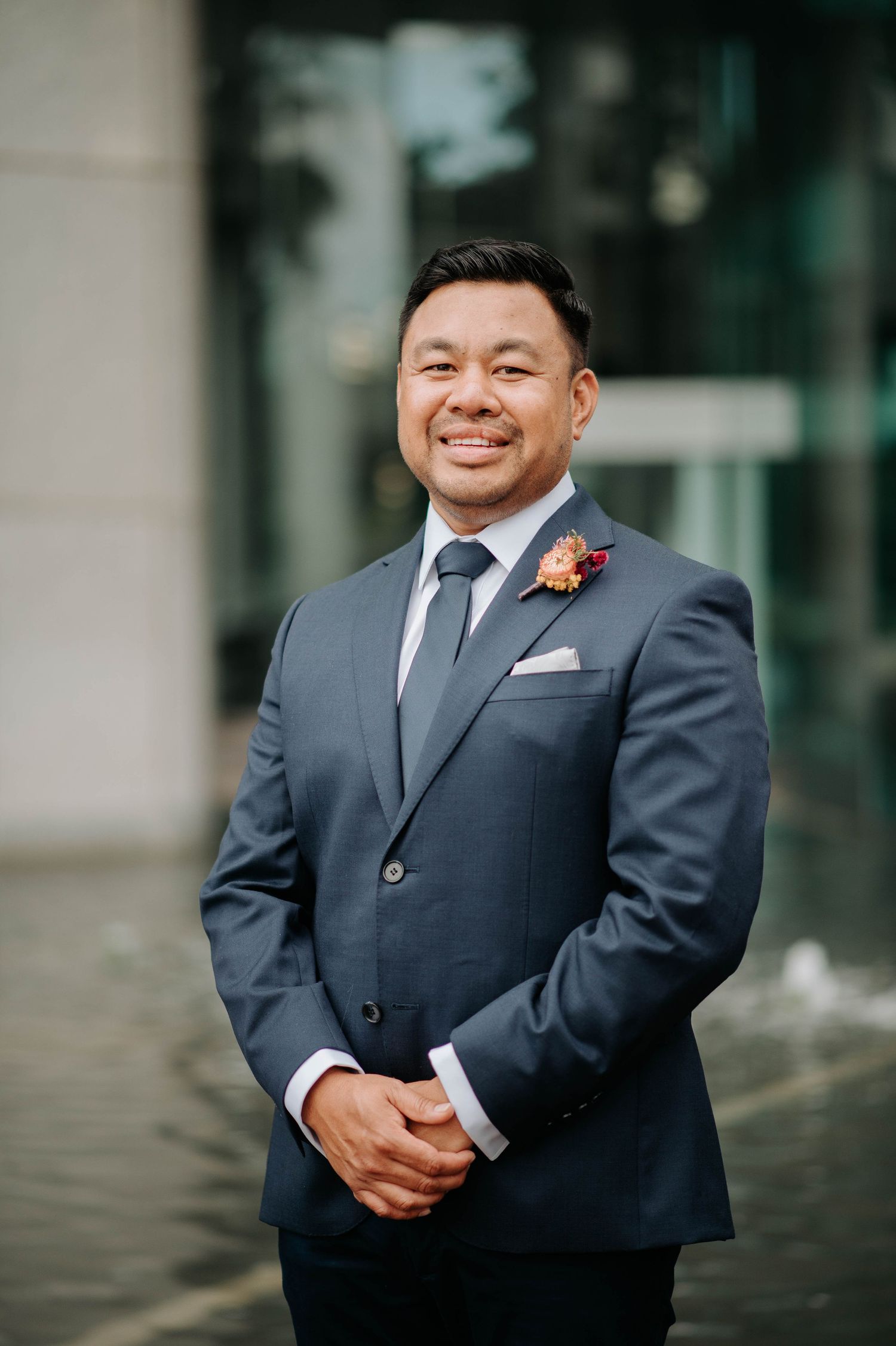 A groom in a grey suit with red boutonniere poses for a formal portrait.