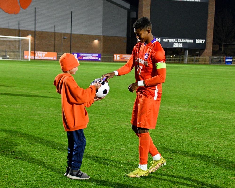 Soccer player in orange uniform interacts with young fan holding a ball during a nighttime training session.