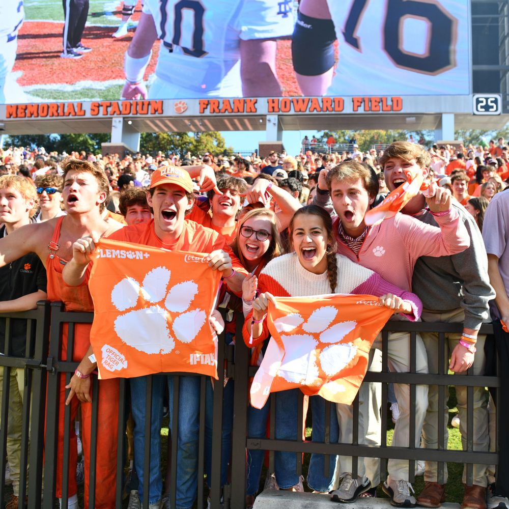 Energetic crowd of Clemson fans cheering with orange tiger paw flags at a football game.