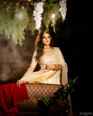 Artistic bridal portrait in cream attire seated under hanging white flowers in moody lighting.