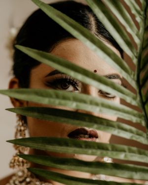 Artistic beauty shot through palm fronds showing detailed eye makeup and jewelry.