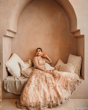 A model in an ornate pink lehenga poses elegantly in an arched alcove with cream cushions.
