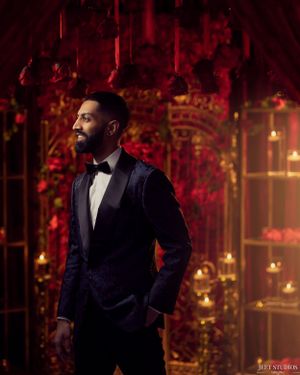 Man in navy velvet tuxedo poses in elegant red and gold setting.