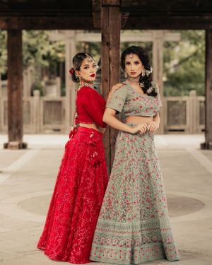 Two models pose in traditional lehengas, one in red and one in grey, under wooden pillars.
