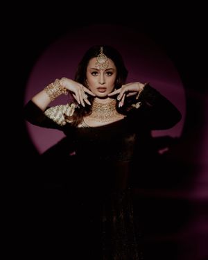 Dramatic portrait in black outfit with traditional Indian jewelry against purple backdrop.