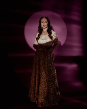 Model in burgundy velvet lehenga poses against purple backlit background in dramatic studio portrait.