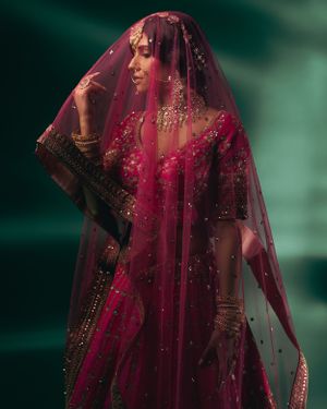 Model in sheer red veil with gold embellishments against emerald green background.