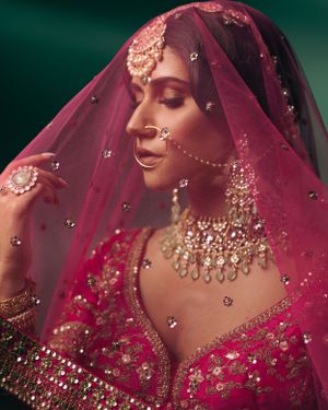 Traditional Indian bride wearing ornate jewelry and burgundy veil with delicate embellishments against dark green background.