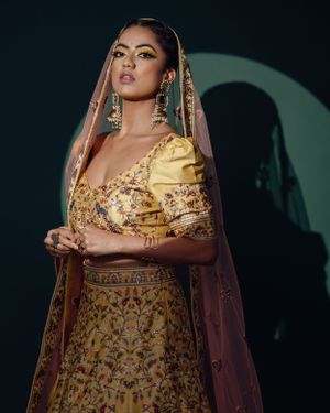 Model wearing yellow and gold embroidered lehenga with sheer dupatta in dramatic studio lighting.