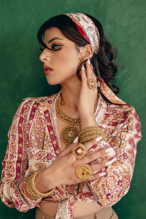Model wearing ornate Indian-inspired jewelry and printed blouse poses against green backdrop in vintage fashion photoshoot.