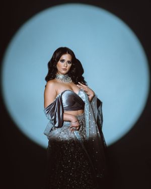Fashion model in silver crop top and glittering black skirt poses against blue circular backdrop.