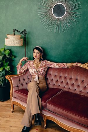 Vintage-inspired fashion shoot with model on velvet sofa featuring retro decor and sunburst mirror.