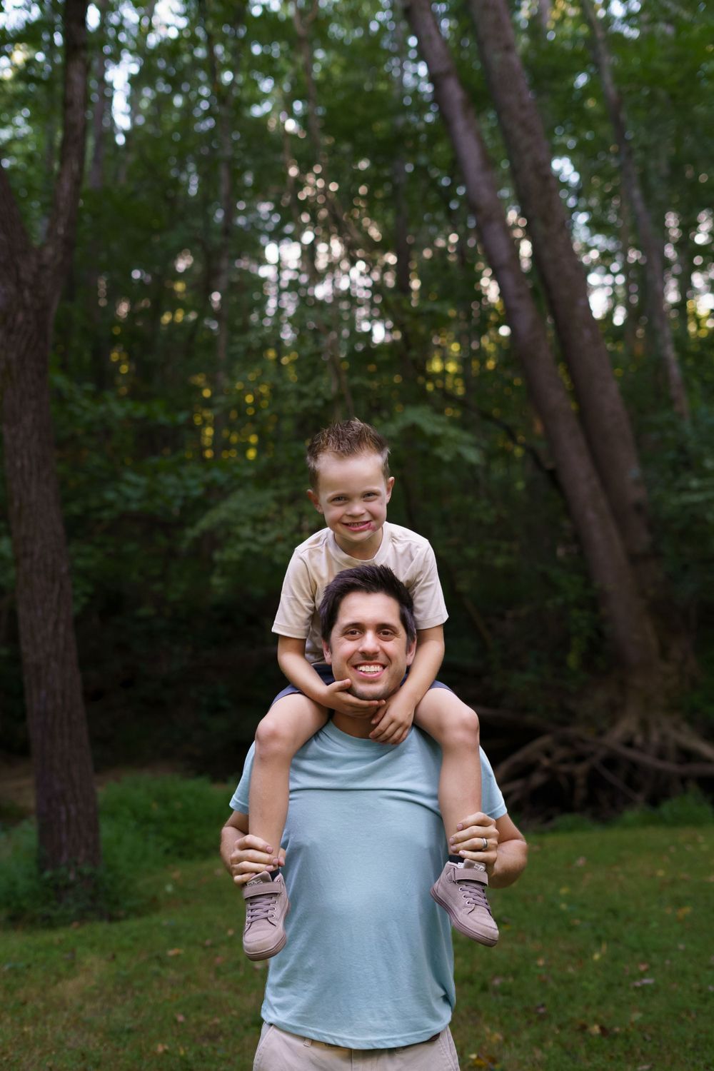 Dad carrying his son on his shoulders through a wooded spot during a family session, captured in a heartfelt moment by Brandy Lee Aldrich.