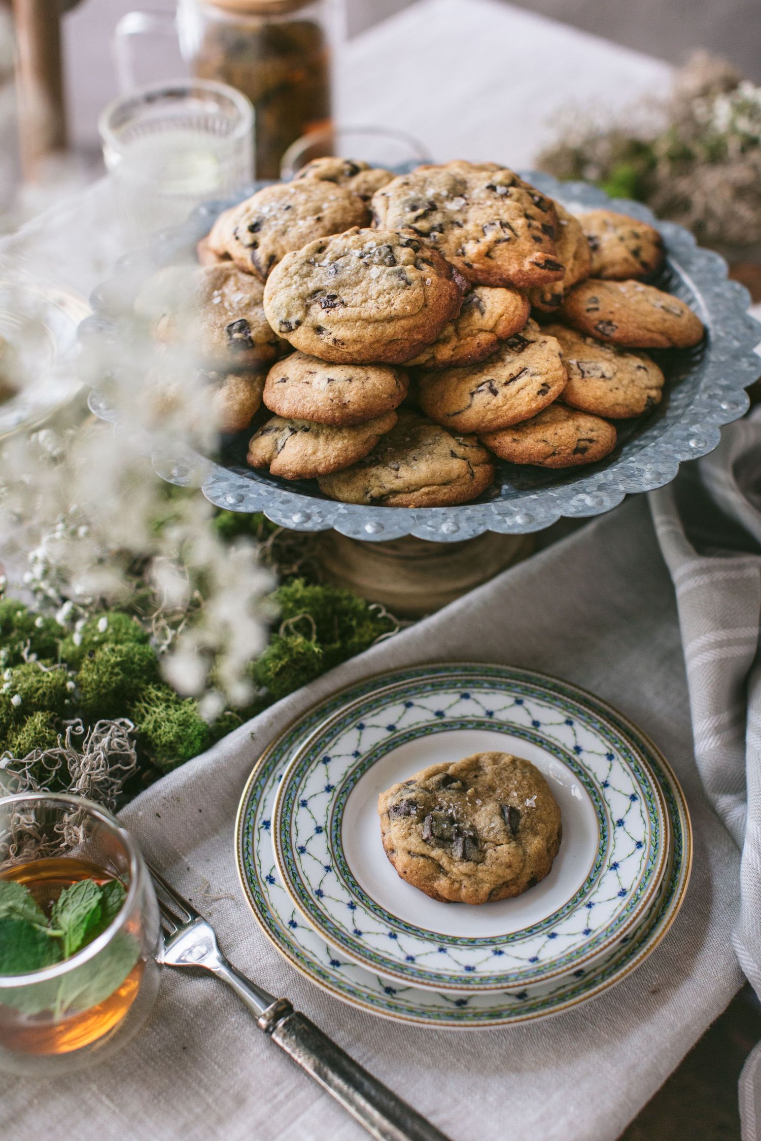 Elegant table setting with chocolate chip cookies on a vintage cake stand, decorated with wildflowers and a cup of herbal tea.