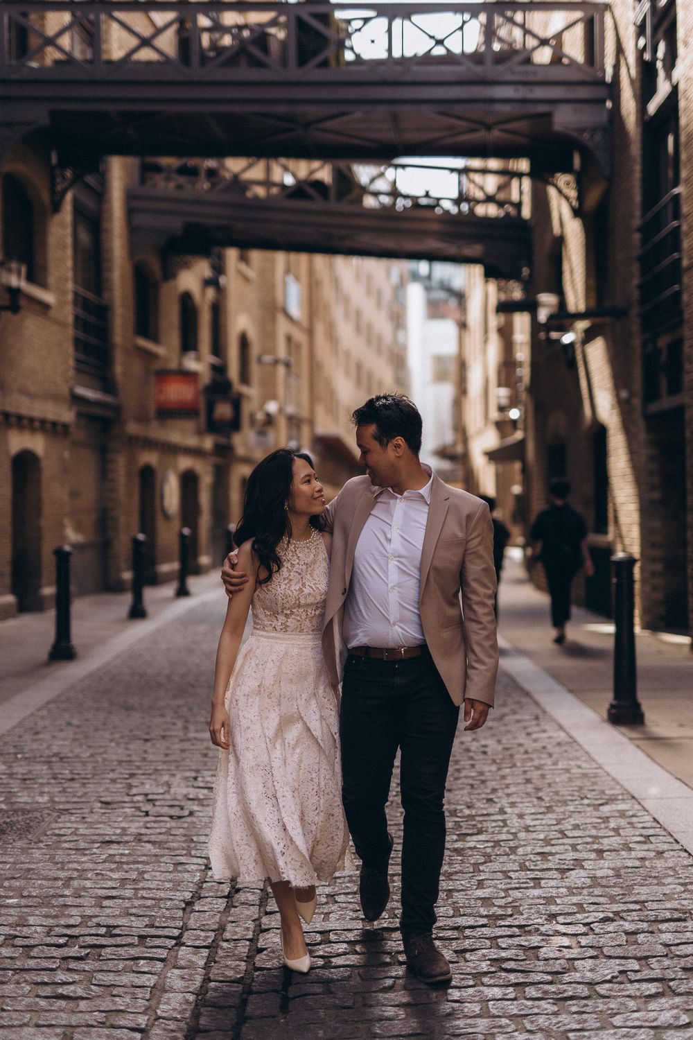 Couple walking together on a cobblestone street with historic bridges overhead.