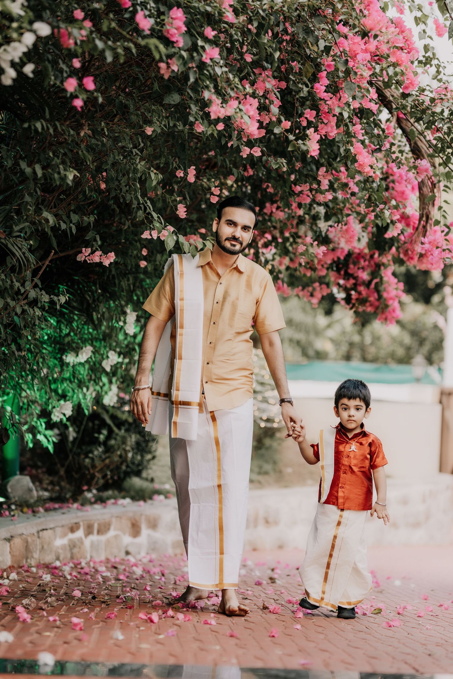 Family in traditional Indian clothing walking together on brick path under pink flowering trees.