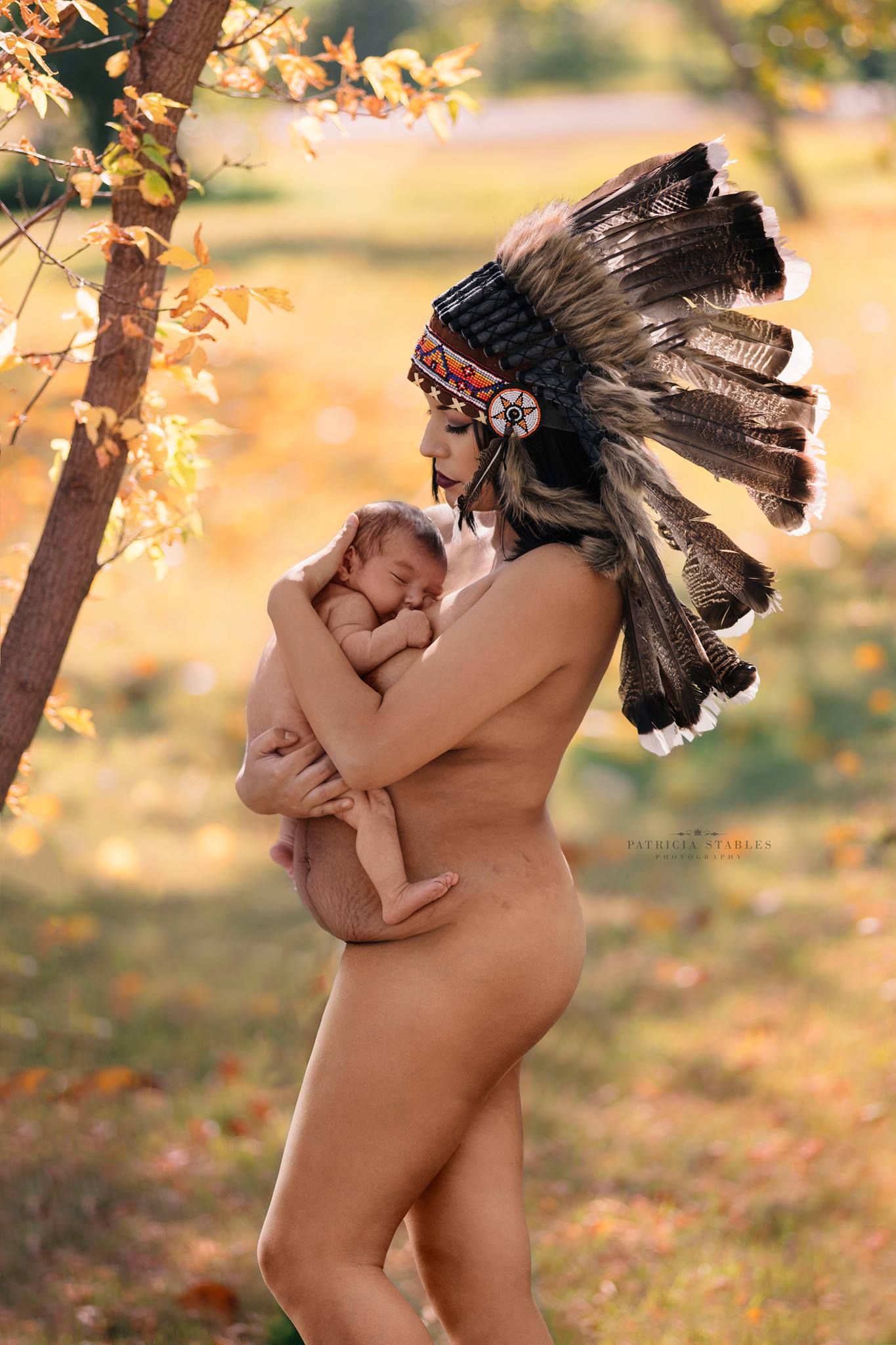Artistic maternity photo featuring traditional Native American headdress in golden autumn setting.